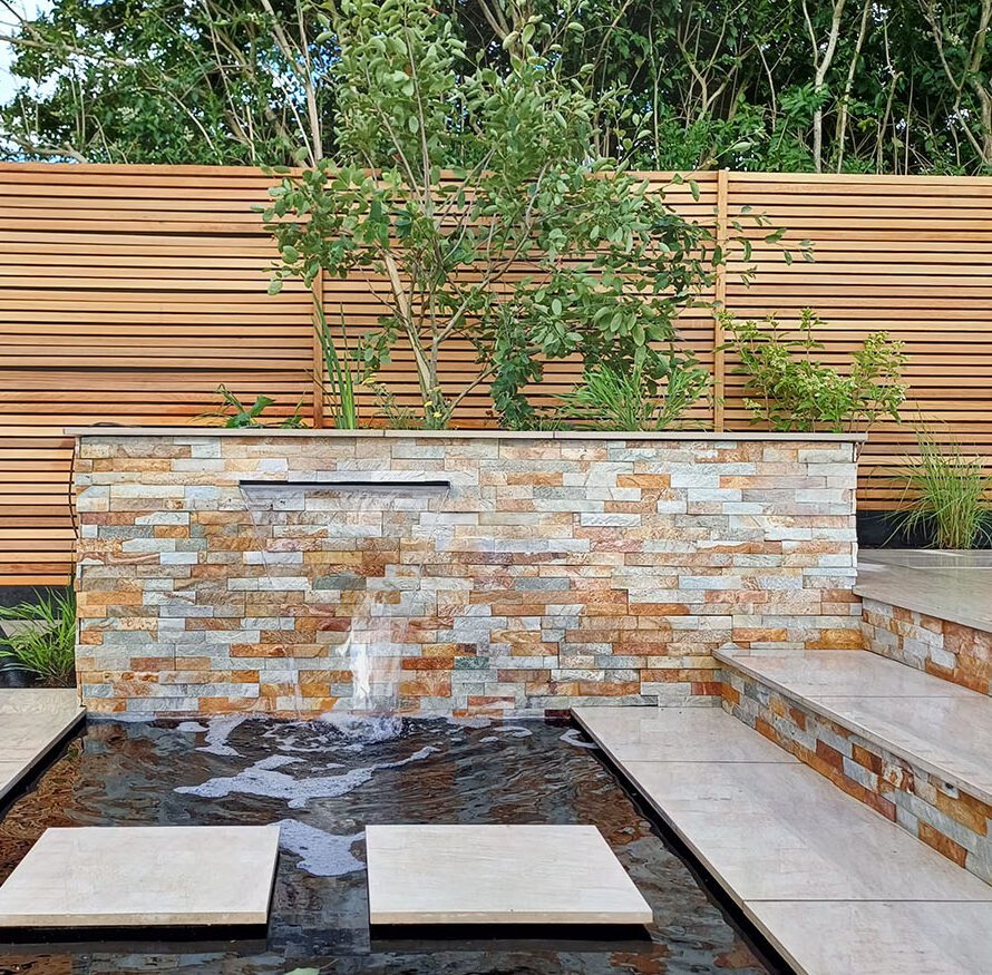Contemporary countryside garden design featuring a striking central planting bed with a sculptural multistem tree, richly textured herbaceous planting, and a sleek blade water feature. Modern rural landscape combining naturalistic planting design with elegant architectural detailing for timeless outdoor style.