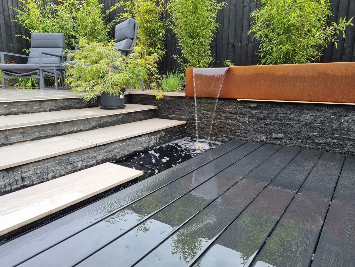 Contemporary garden design featuring a sleek central water feature set against dark timber decking, elegant porcelain paving, and sculptural corten steel features and planters, including a distinctive coffin shaped planter. Modern architectural garden combining strong materials, clean lines, and striking design contrasts for a bold outdoor aesthetic