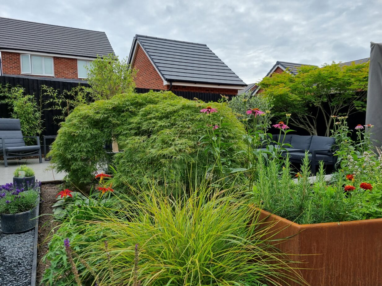 Contemporary garden design featuring a sleek central water feature set against dark timber decking, elegant porcelain paving, and sculptural corten steel features and planters, including a distinctive coffin shaped planter. Modern architectural garden combining strong materials, clean lines, and striking design contrasts for a bold outdoor aesthetic