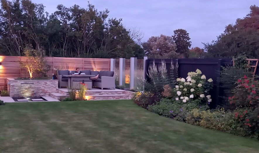 Contemporary countryside garden design featuring a striking central planting bed with a sculptural multistem tree, richly textured herbaceous planting, and a sleek blade water feature. Modern rural landscape combining naturalistic planting design with elegant architectural detailing for timeless outdoor style.
