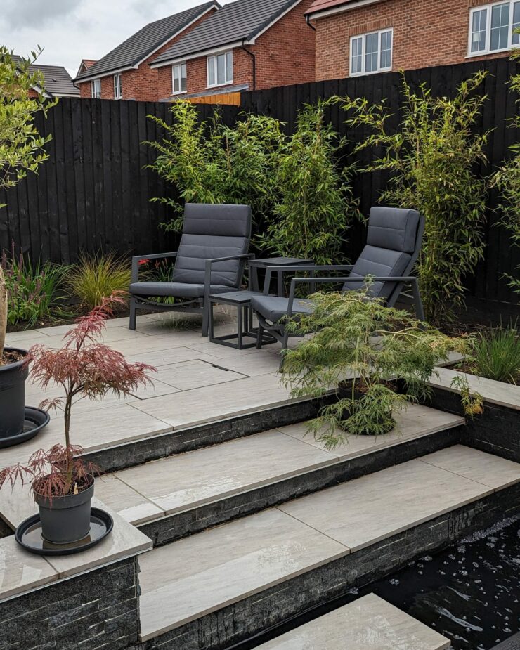 Contemporary garden design featuring a sleek central water feature set against dark timber decking, elegant porcelain paving, and sculptural corten steel features and planters, including a distinctive coffin shaped planter. Modern architectural garden combining strong materials, clean lines, and striking design contrasts for a bold outdoor aesthetic