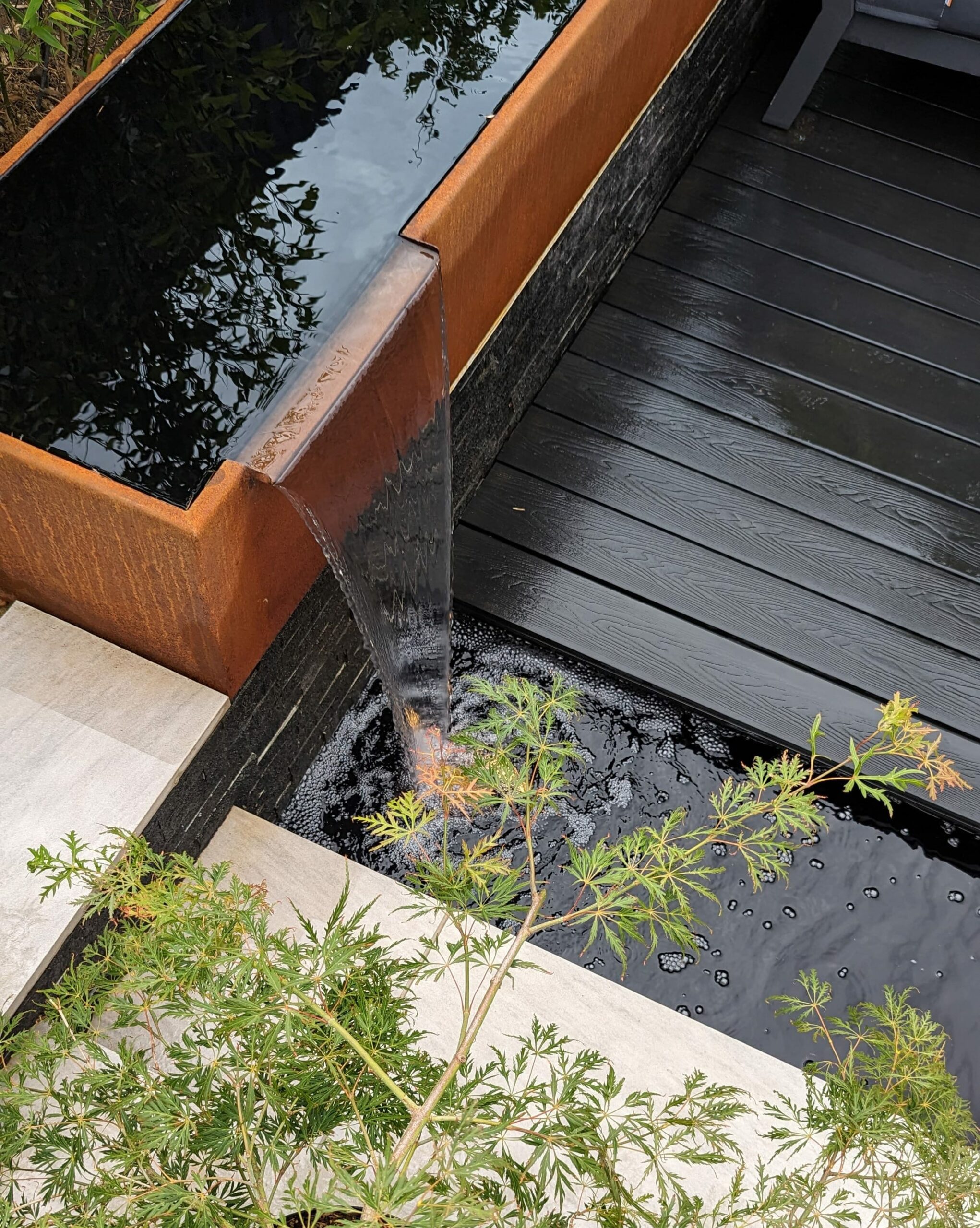 Contemporary garden design featuring a sleek central water feature set against dark timber decking, elegant porcelain paving, and sculptural corten steel features and planters, including a distinctive coffin shaped planter. Modern architectural garden combining strong materials, clean lines, and striking design contrasts for a bold outdoor aesthetic