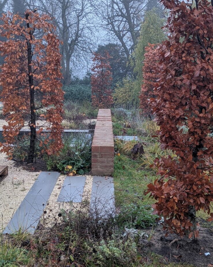 Contemporary new build garden set in a woodland landscape with extensive matrix planting and a sculptural corten steel water feature. Modern naturalistic garden design featuring layered perennial planting, ornamental grasses, and native woodland plants blending seamlessly with the surrounding trees. A tranquil, sustainable outdoor space combining modern architecture with organic landscape design.