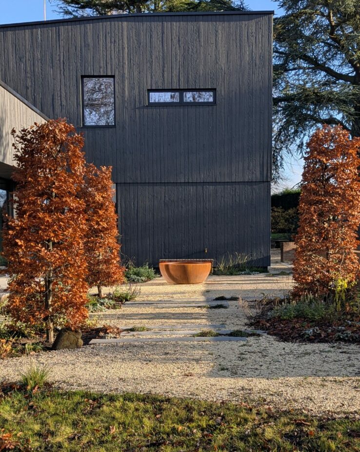 Contemporary new build garden set in a woodland landscape with extensive matrix planting and a sculptural corten steel water feature. Modern naturalistic garden design featuring layered perennial planting, ornamental grasses, and native woodland plants blending seamlessly with the surrounding trees. A tranquil, sustainable outdoor space combining modern architecture with organic landscape design.